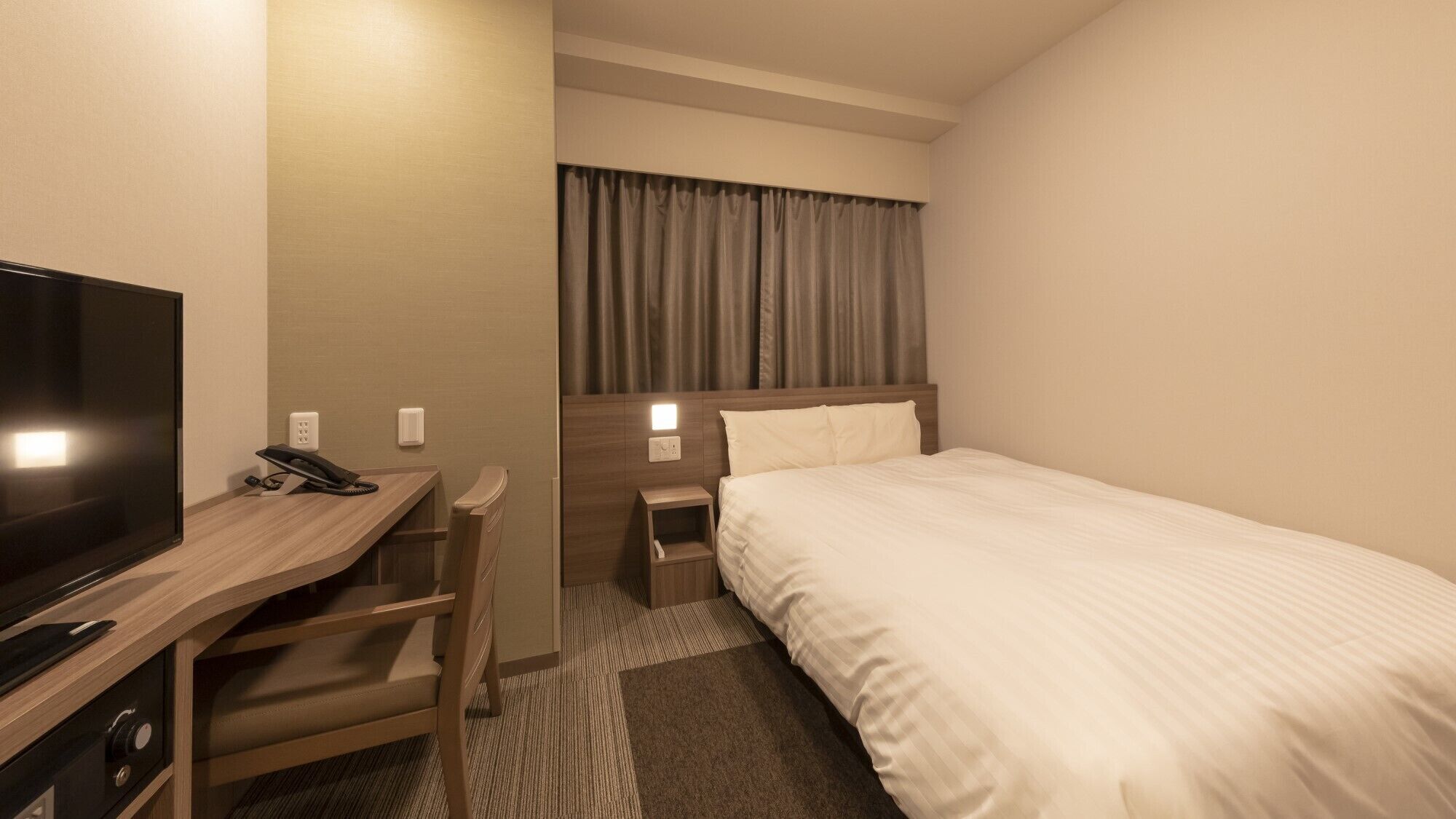 Een hotelkamer met een bed, een bureau met televisie, een telefoon en een nachtkastje met lamp.