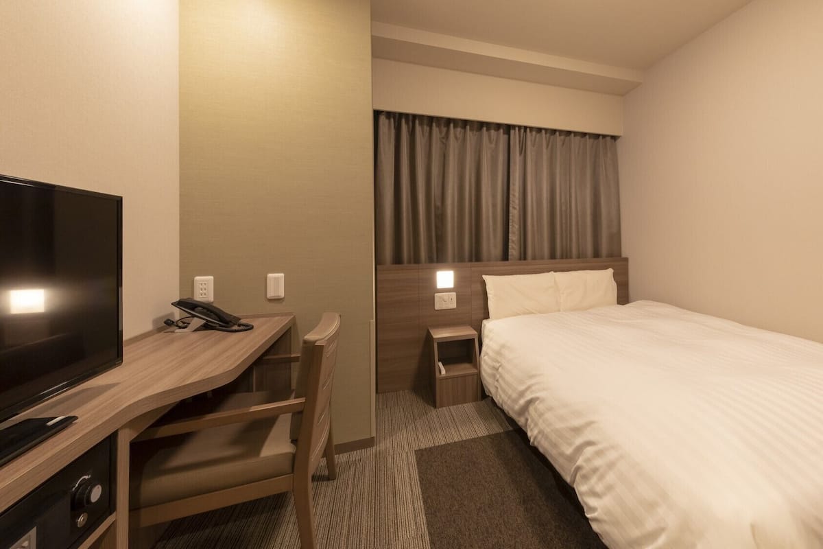 A hotel room with a bed, a desk with a television, a phone, and a nightstand with a lamp.
