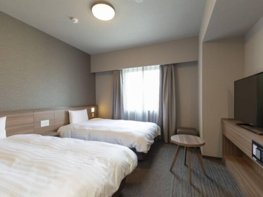 A hotel room with two beds, a TV, a small table, and a window with curtains.