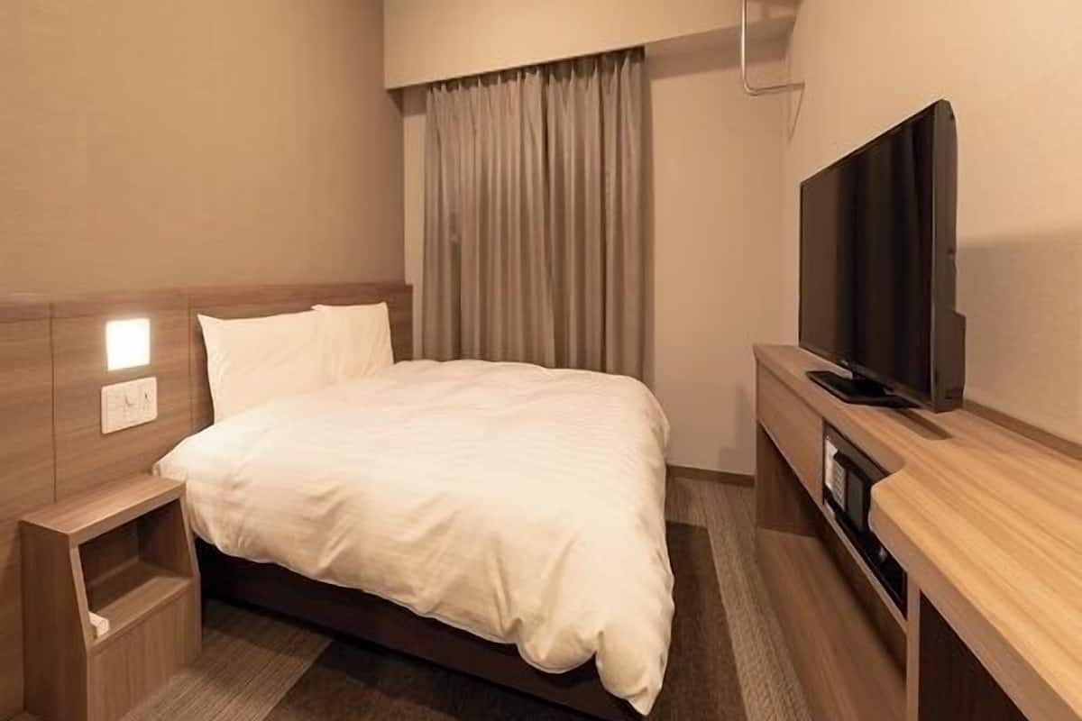 A hotel room with a bed, a TV, a bedside table, and a window with curtains.