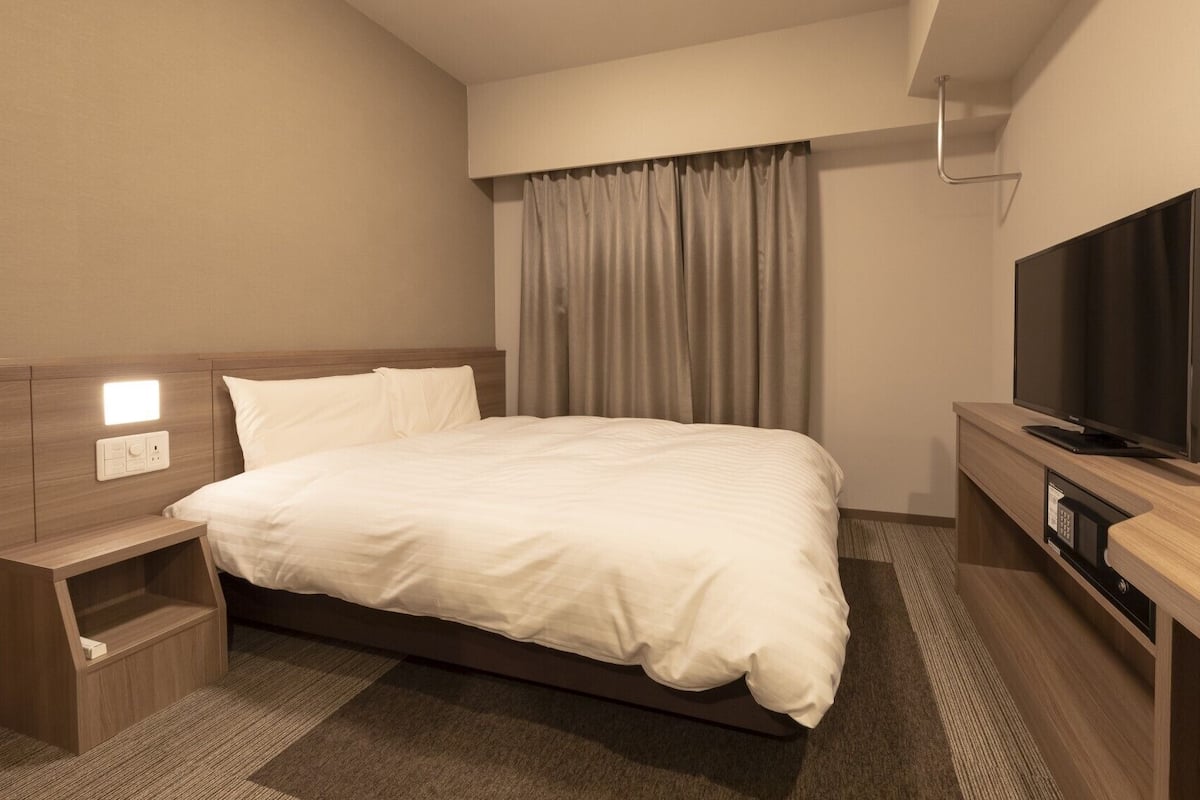 A hotel room with a bed, bedside table, TV, and a control panel on the wall.