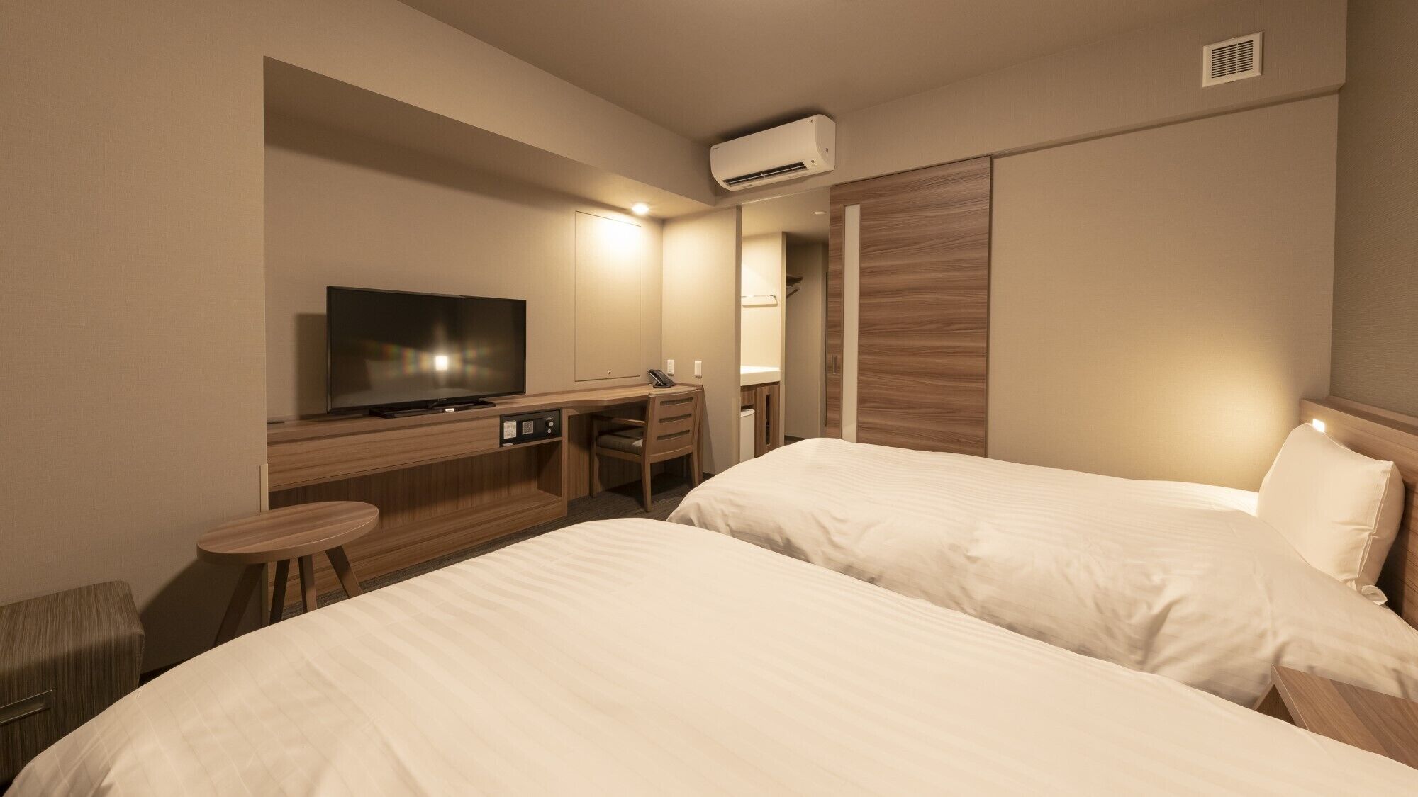Een hotelkamer met twee bedden, een bureau met een televisie, een stoel en een airconditioning.