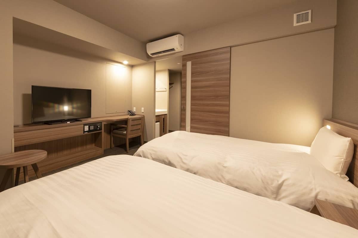 A hotel room with two beds, a desk with a TV, a chair, and an air conditioning unit.