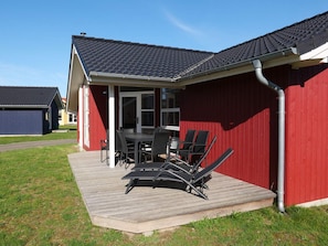 Eine überdachte Terrasse mit Tisch und Stühlen, eine Holzterrasse und ein rotes Gebäude mit dunklem Dach.