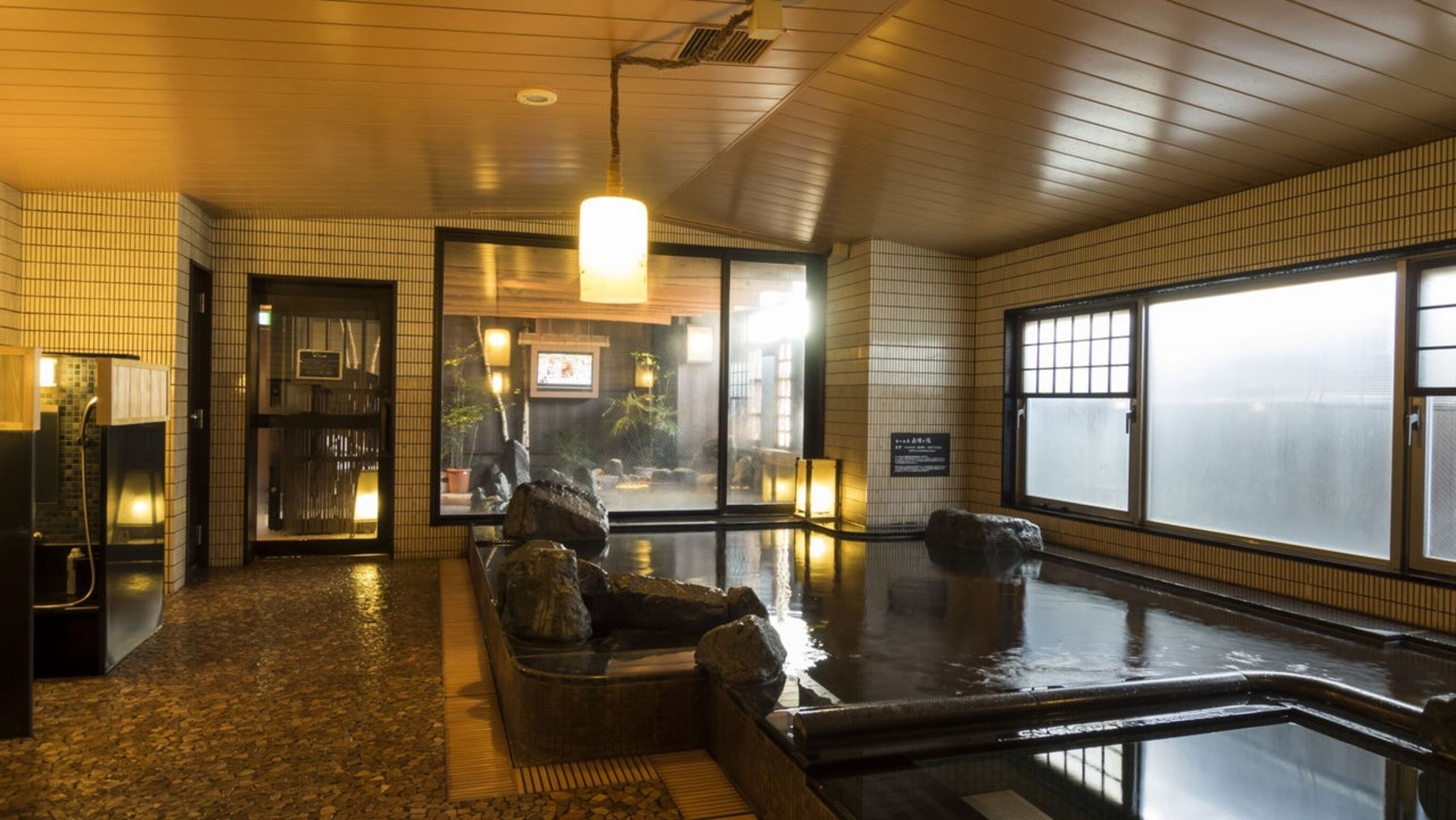 Un onsen japonés tradicional con interior de madera, un baño de aguas termales y vistas a través de un ventanal grande.