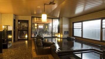 Un onsen japonés tradicional con interior de madera, un baño de aguas termales y vistas a través de un ventanal grande.