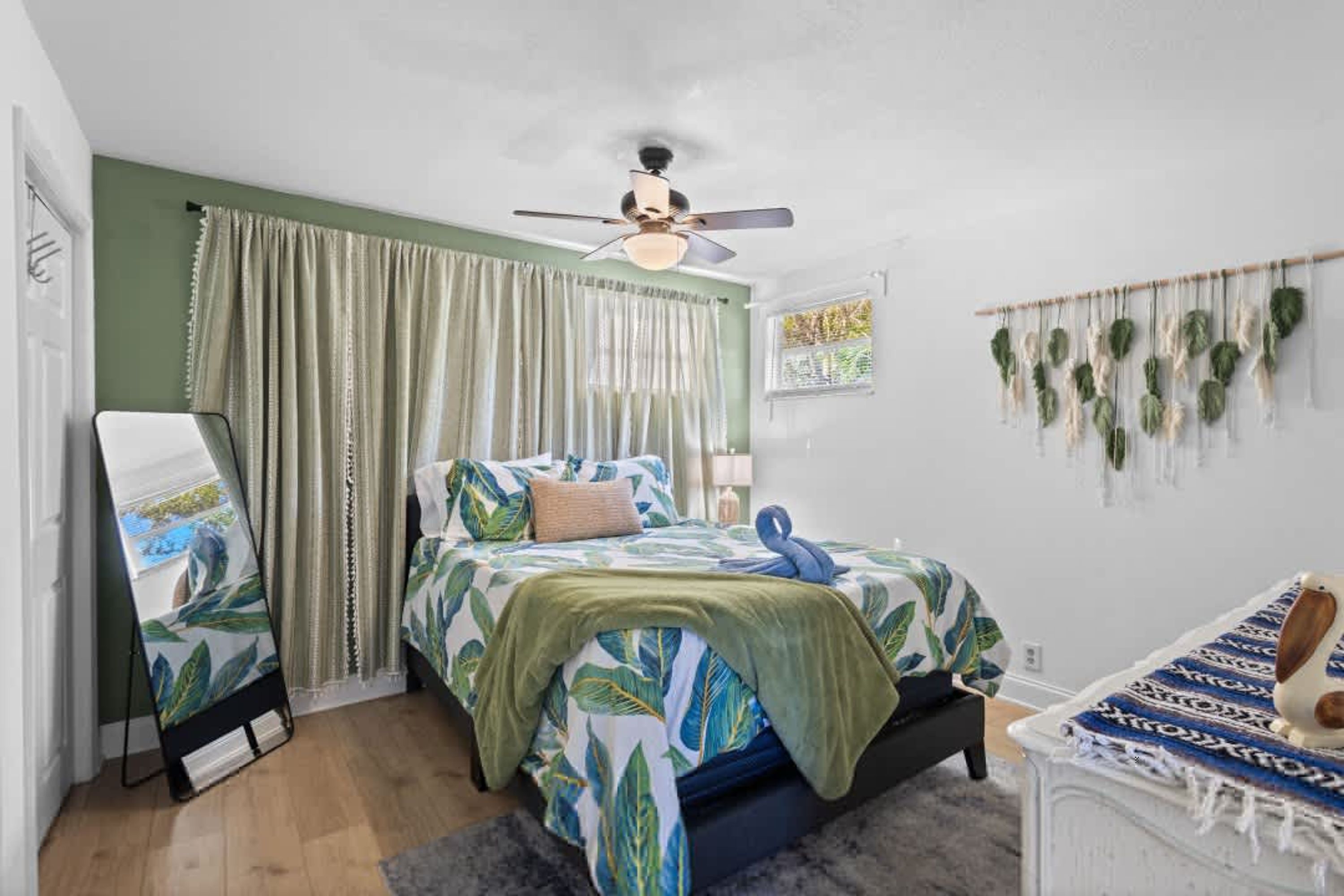 A bedroom with a bed, a ceiling fan, curtains, a wall-mounted plant, and a small table with a lamp.
