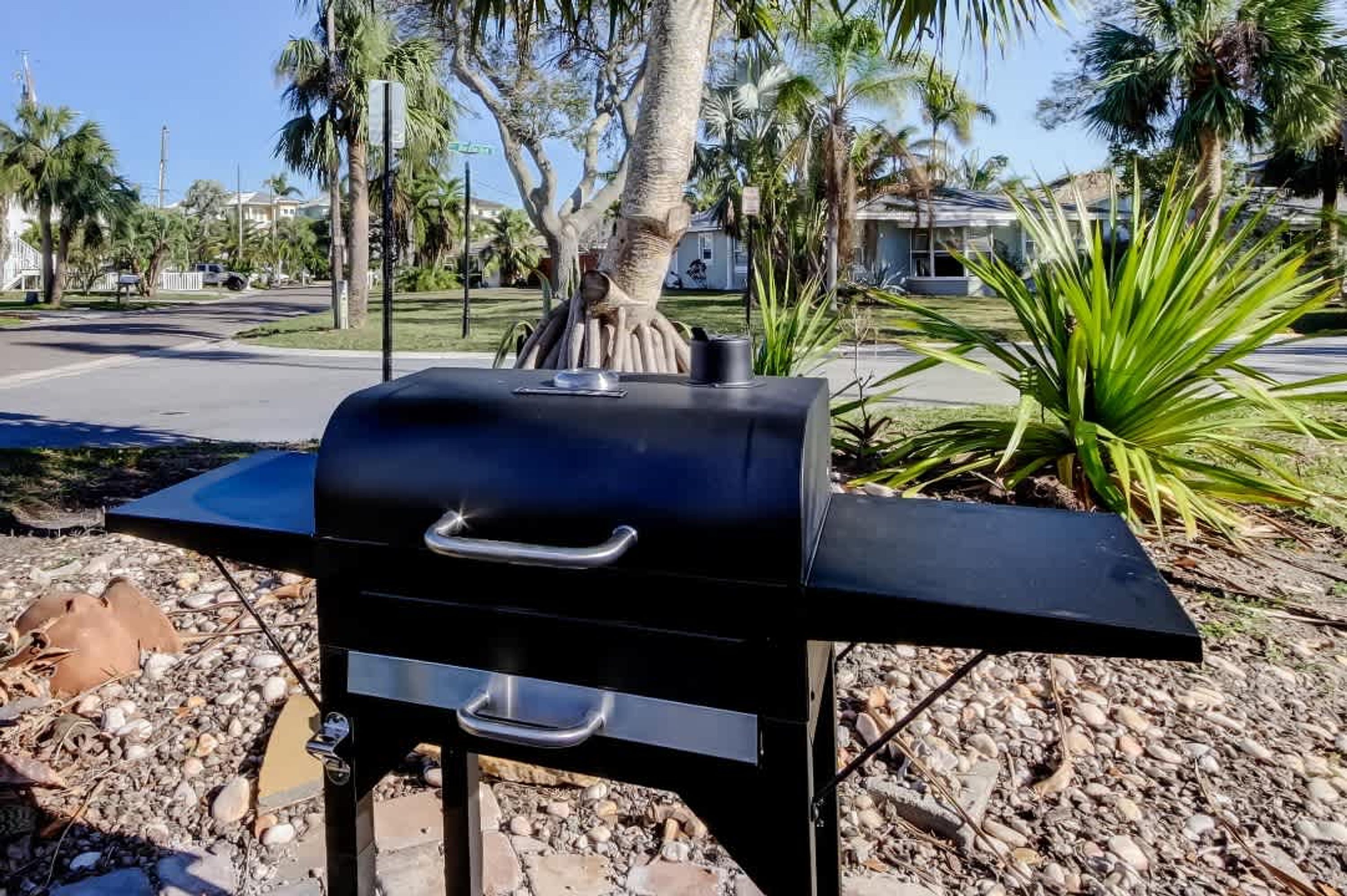A black barbecue grill with a handle and a vent, set on a gravel surface with palm trees and houses in the background.