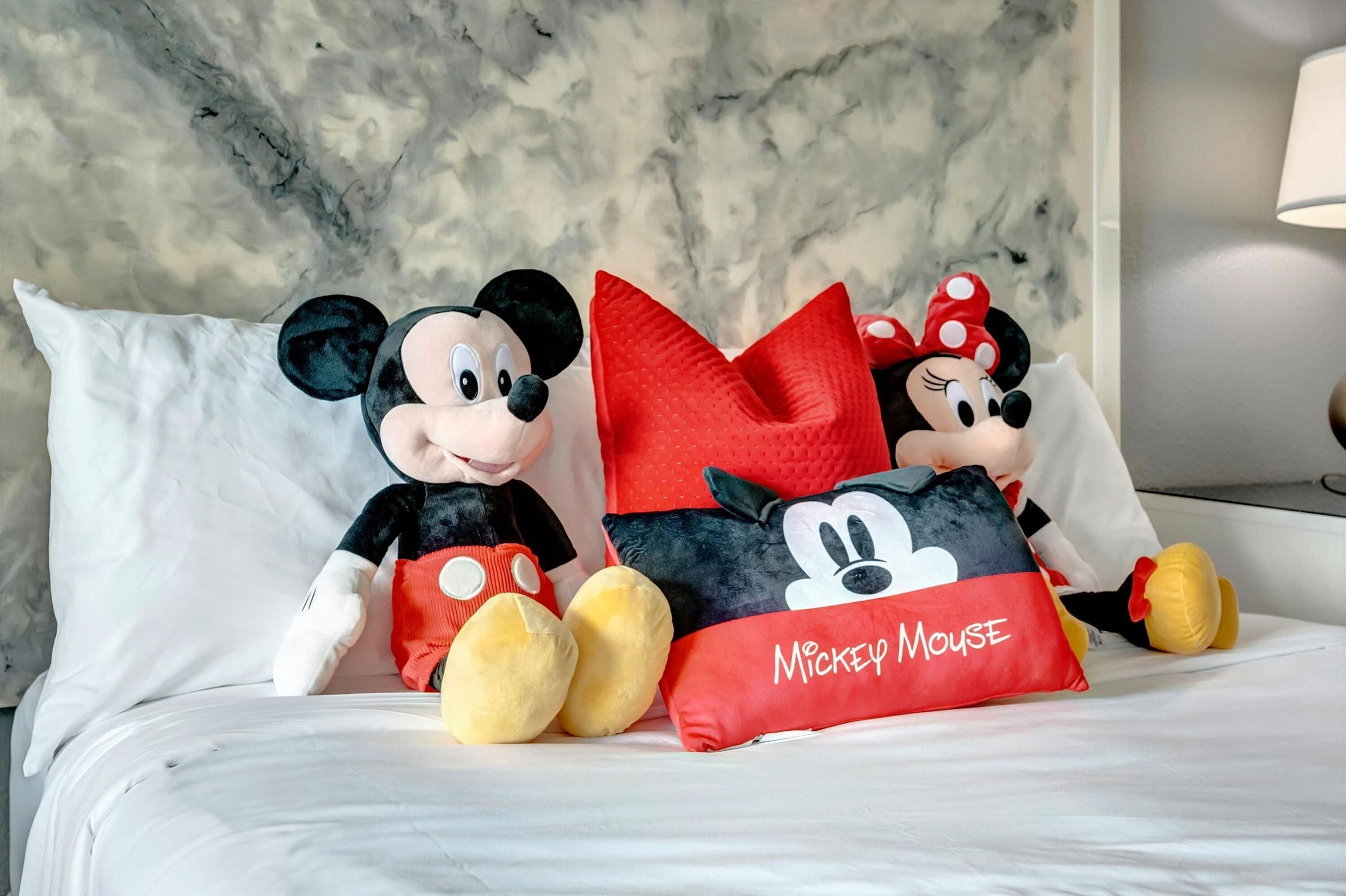 Peluche de Mickey y Minnie Mouse sobre una cama, con un muro de diseño de mármol y una lámpara al fondo.