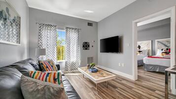A modern living room with a grey couch, a wooden coffee table, a TV mounted on the wall, and a bed in the background.