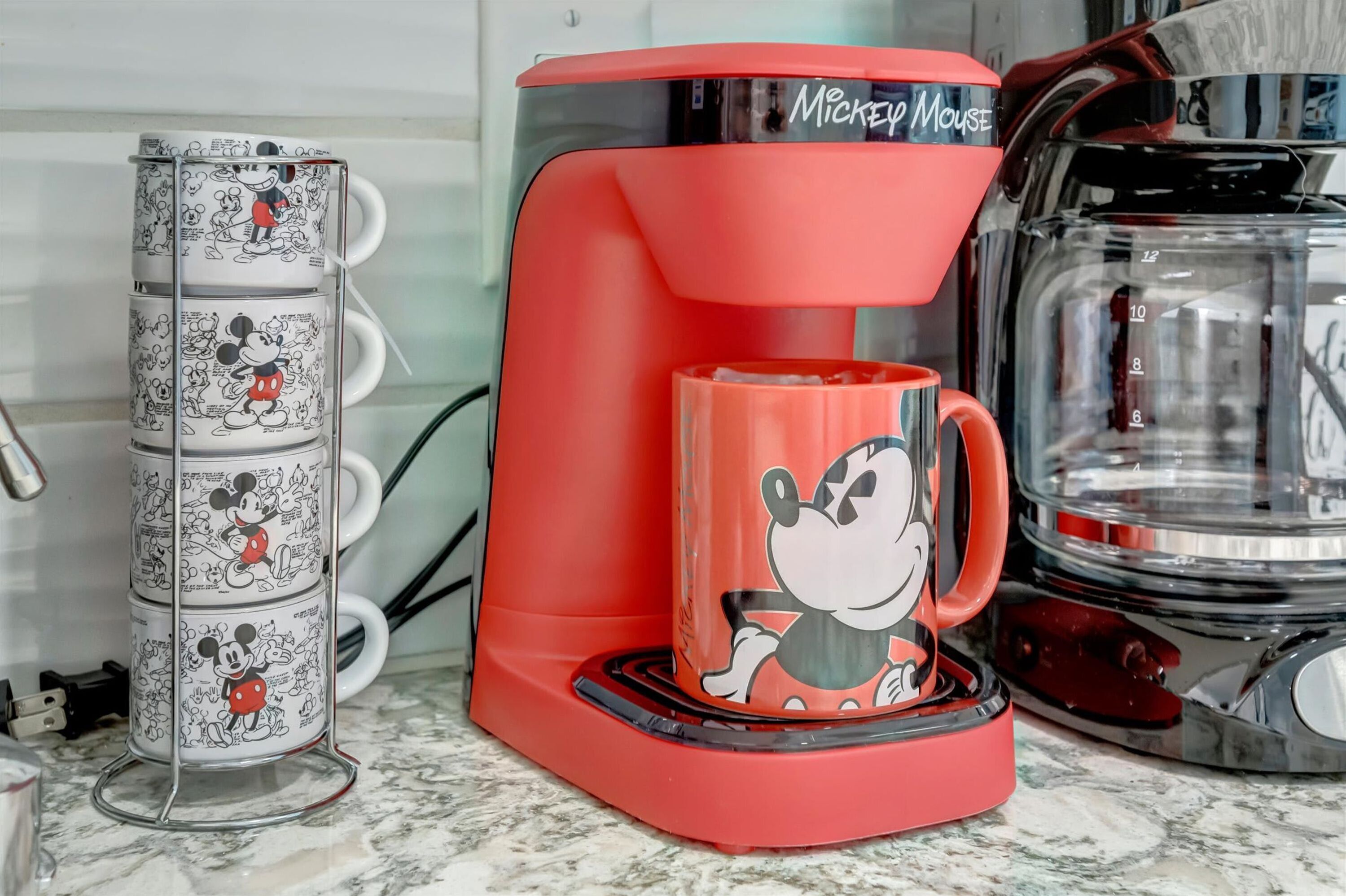Une machine à café accompagnée d’une tasse à l’effigie de Mickey Mouse, un porte-tasses avec des tasses à l’effigie de Mickey Mouse et une cafetière.