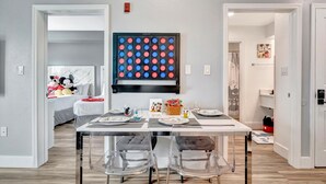 Un coin repas avec une table dressée pour quatre personnes, des chaises transparentes, une œuvre d’art accrochée au mur et, au fond, un lit avec des peluches.