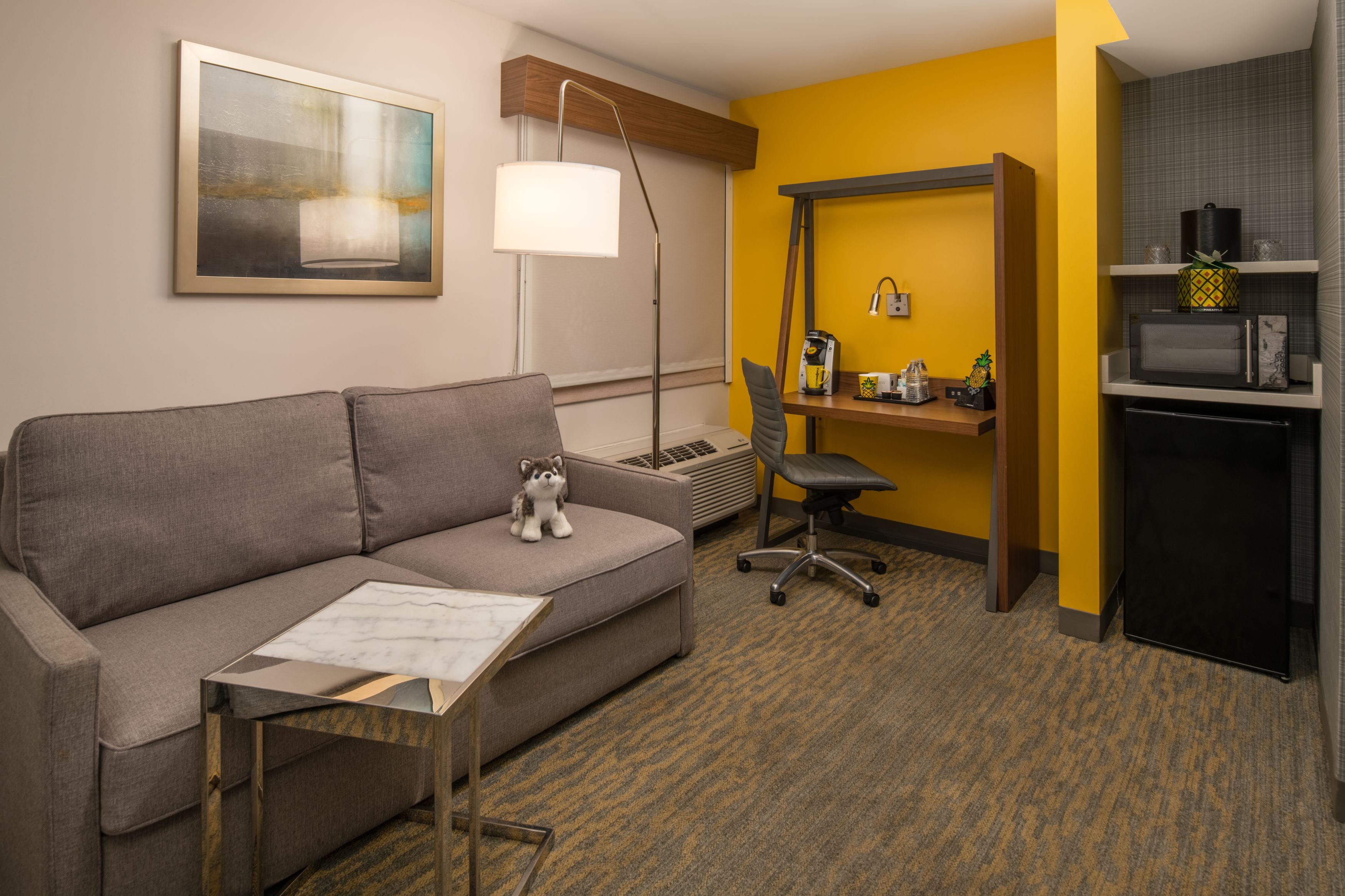 A hotel room with a gray sofa, a small table, a lamp, a kitchenette, and a painting on the wall.
