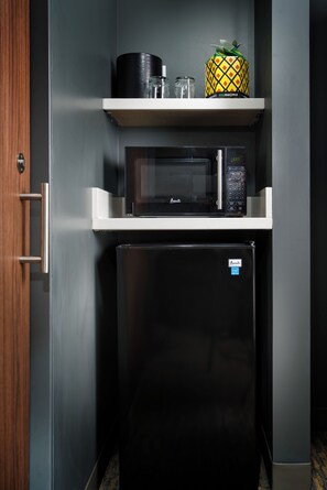 Una kitchenette compacta con microondas, un refrigerador pequeño y una estantería con un recipiente con forma de piña.