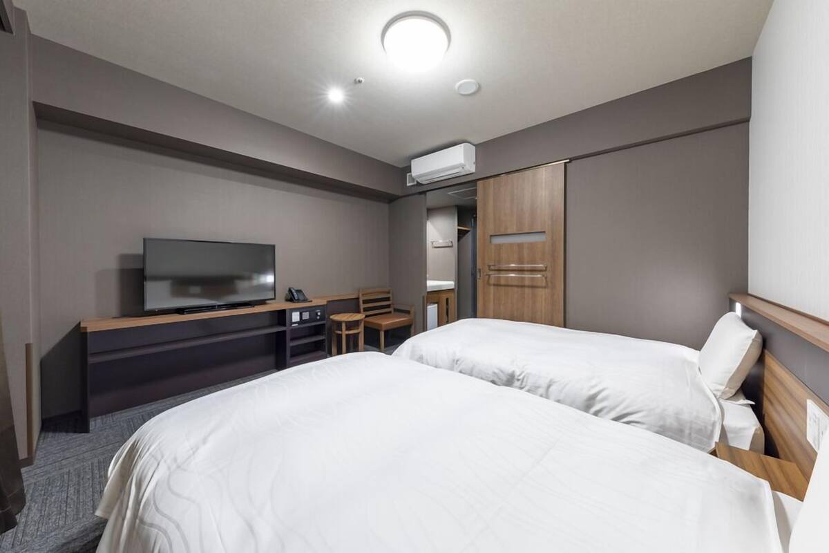 A hotel room with two beds, a desk with a TV, a chair, a wardrobe, and an air conditioning unit.