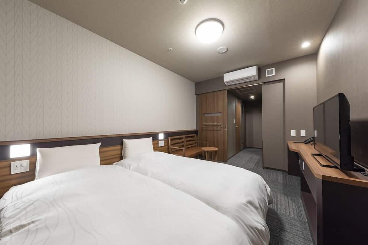 A hotel room with two beds, a desk, a television, and an air conditioning unit.