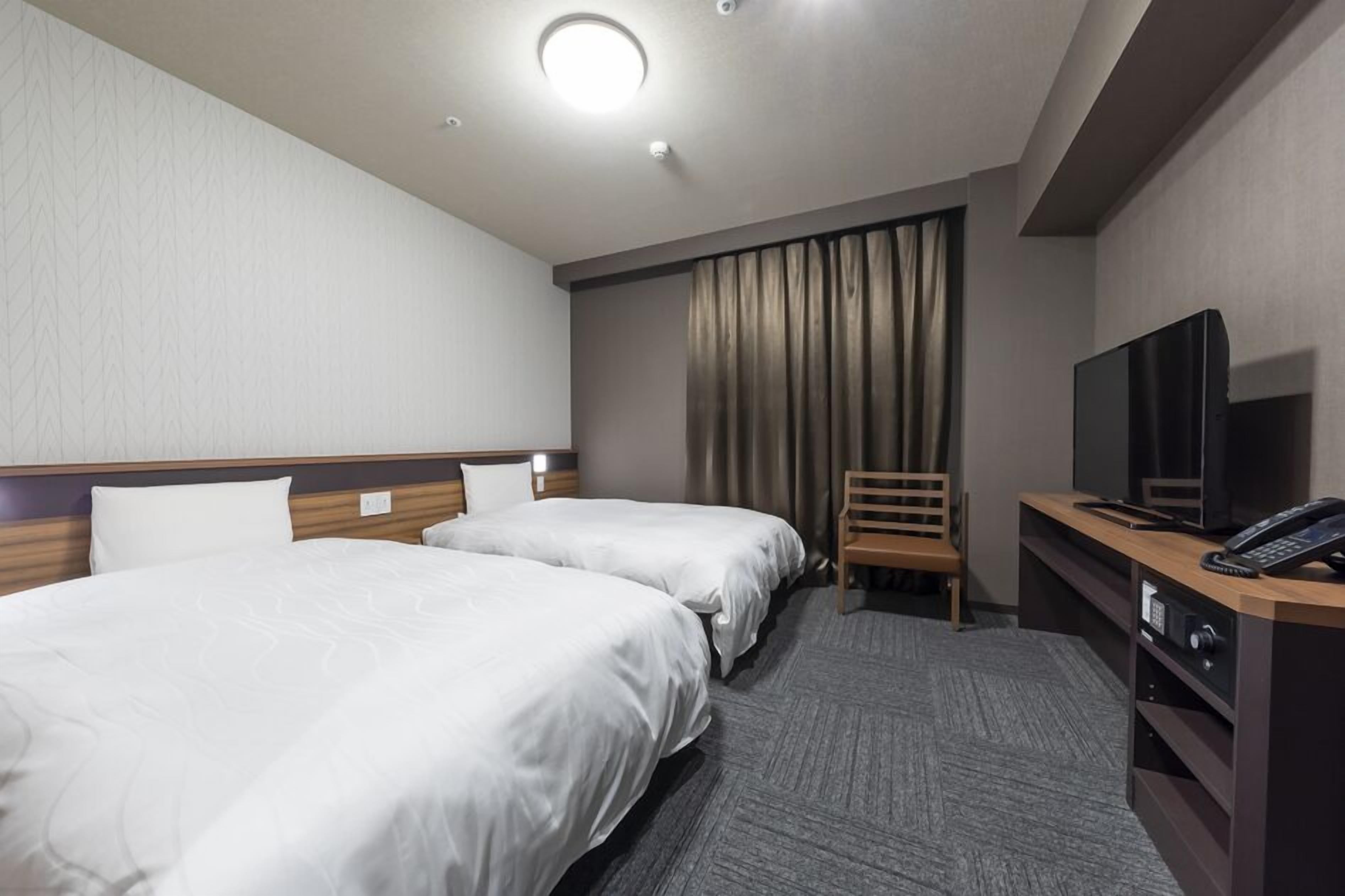 Une chambre d’hôtel avec deux lits, un bureau équipé d’une télévision et d’un téléphone, et une fenêtre avec des rideaux.