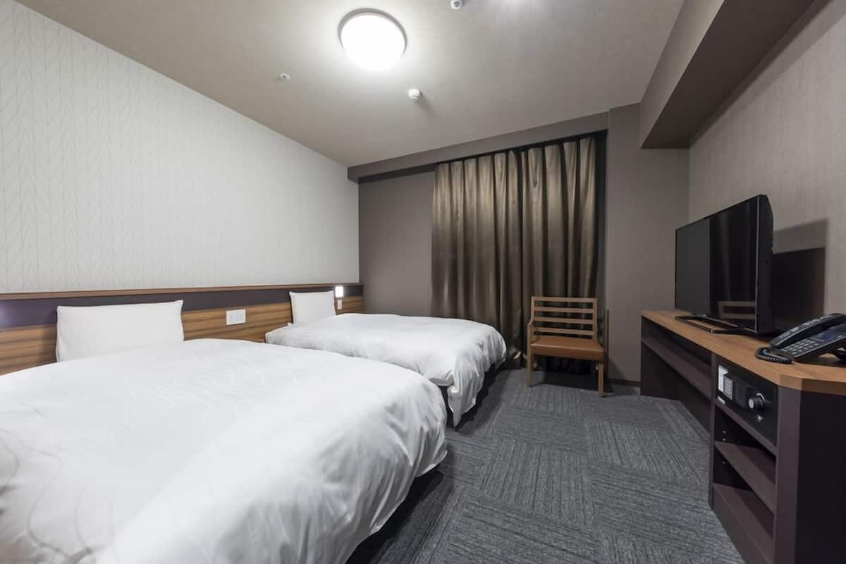 A hotel room with two beds, a desk with a TV and phone, and a window with curtains.