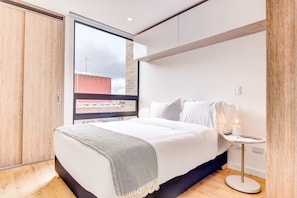 Ein modernes Schlafzimmer mit großem Fenster, einem Bett mit weißer Bettwäsche, einem Nachttisch mit Lampe und Holzboden.