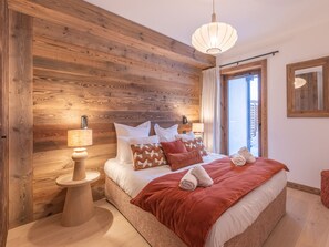A bedroom with a wooden wall, a bed with pillows, a nightstand with a lamp, and a window with curtains. - Apartment in the heart of the Alps - 4 guests (Huez)