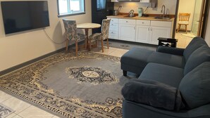 A living room with a sofa, a TV, a dining table with chairs, a kitchen area with cabinets and appliances, and a patterned rug.