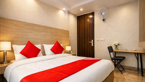 A hotel room with a bed, bedside tables, a desk, a chair, a fan, and a door. - Hotel Vindhya Inn - Delhi Airport (New Delhi)