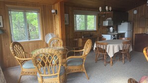A wooden cabin interior with wicker furniture, a dining table, and a kitchen area. - Peaceful Kawartha Lakes Cottage | Kayak, SUP & Stunning Sunsets (North Kawartha)