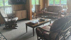 A cozy cabin interior with a wooden ceiling, a television mounted on the wall, a rocking chair, a sofa, and a coffee table with books. - Peaceful Kawartha Lakes Cottage | Kayak, SUP & Stunning Sunsets (North Kawartha)
