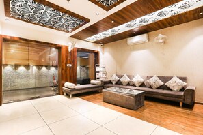 A lobby with a sofa set, a glass coffee table, a patterned ceiling, and a wooden floor.