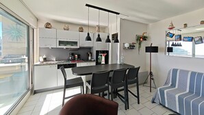 A modern kitchen with white cabinets, a dining area with a black table and chairs, a sofa with striped cushions, and a large window with a view. - Stunning T2 sea view - 4 guests (Canet-en-Roussillon)