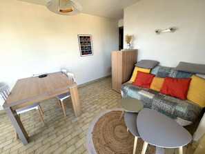 A living area with a wooden dining table, a sofa with patterned cushions, a small round table, and a woven rug. - Sea view apartment, private parking - 4 guests (Argelès-sur-Mer)