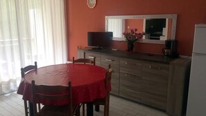 Un coin repas avec une table et des chaises rouges, un téléviseur posé sur un meuble en bois, un réfrigérateur et une fenêtre avec des rideaux.