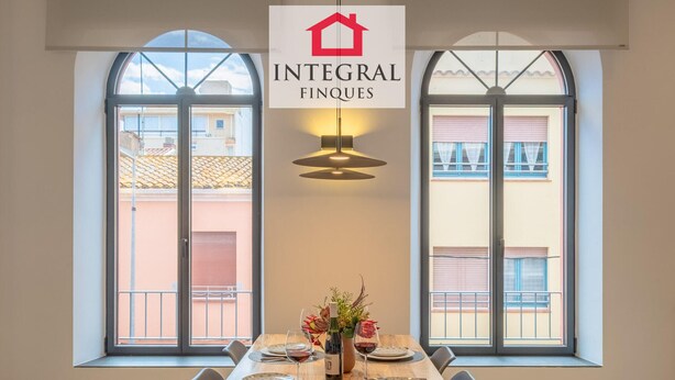 Un comedor con una mesa de madera dispuesta para cuatro personas, dos ventanas grandes con balcones, y un letrero que dice 'INTEGRAL FINQUES'.