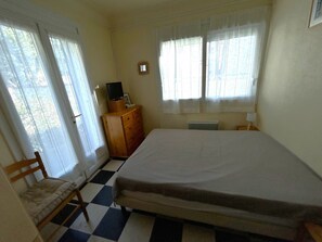Ein Zimmer mit einem Bett, einem Sessel, einer Holzkommode, einem kleinen Tisch mit Lampe und einem Fenster mit Vorhängen.