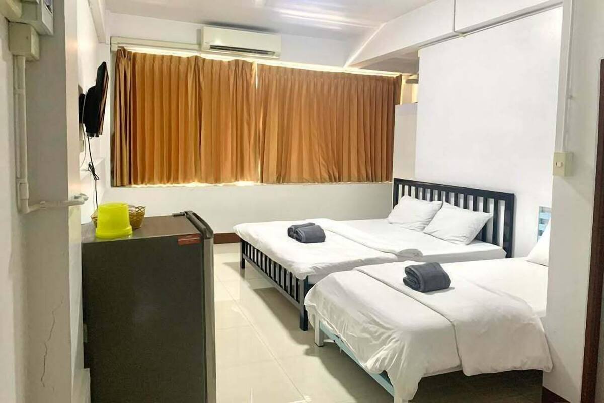 A hotel room with two beds, a desk, a chair, a small table, a window with curtains, and an air conditioning unit.