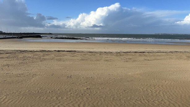 Ein Strand mit felsigem Ufer, blauem Himmel und Gebäuden in der Ferne.