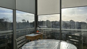 Ein Zimmer mit einem großen Fenster, einem runden Tisch mit gemustertem Tuch, einem kleinen Holznachttisch und einem Stuhl.