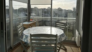 Ein Balkon mit Glastisch und Stühlen, ein kleiner Tisch mit Buch und Glas, und Blick auf Häuser und Bäume.