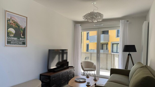 En stue med sofa, sofabord, fjernsyn, stol og en balkon med udsigt til bygninger.