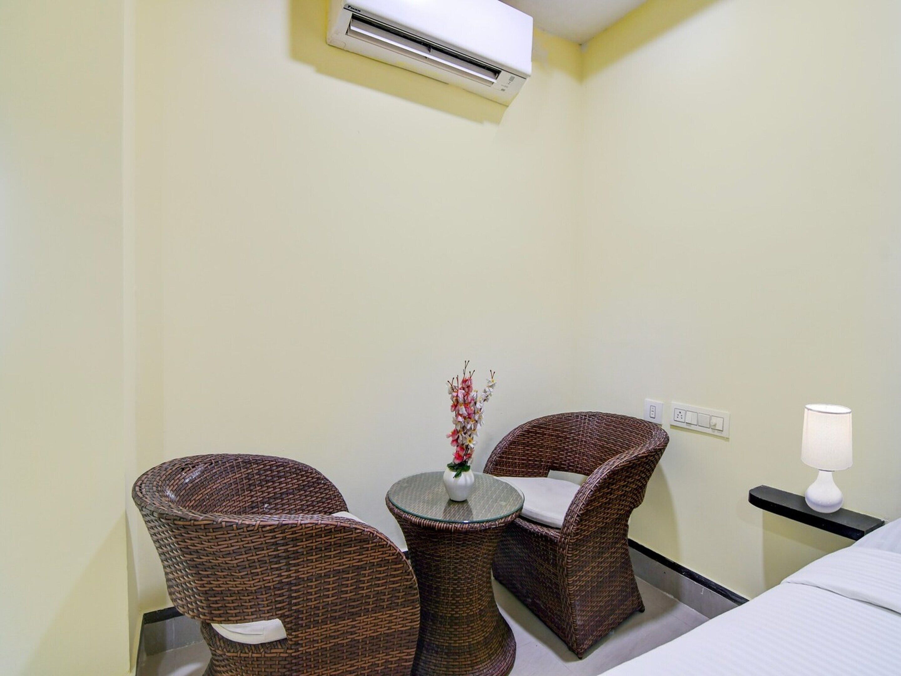 A hotel room with wicker furniture, a small table with a vase, an air conditioning unit, a bedside lamp, and a bed.