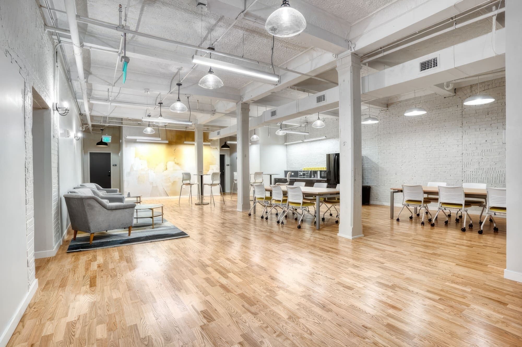 A spacious, modern office with wooden floors, white walls, and exposed ceilings. There are several tables and chairs arranged for meetings, and a large screen for presentations.
