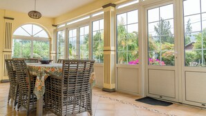 A sunlit room with a table and chairs, large windows, and a view of greenery outside.