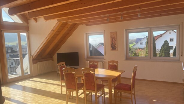 A dining area with a wooden table and chairs, a television, and large windows with a view of houses. - Vacation apartment Loder 108 m2 (Blumberg)