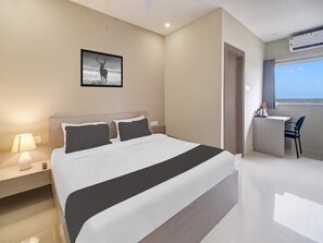 A modern hotel room with a large bed, a desk with a chair, a window with a view, and a wall-mounted air conditioning unit. - Saver Single Cloud Stay (Guwahati)