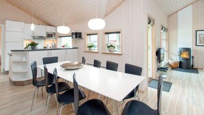 Una cocina moderna con armarios blancos y una isla central, un comedor con mesa blanca y sillas negras, y techo revestido de madera.