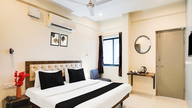 A hotel room with a bed, bedside table, air conditioning unit, ceiling fan, window with curtains, and a door. - Deluxeorient Royale Nagpur (Nagpur)