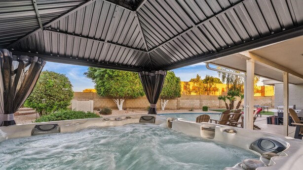 Área de hidromassagem com teto coberto, espaço para sentar ao ar livre e vista para uma piscina e vegetação.