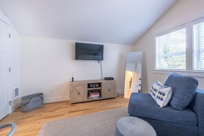 Ein Zimmer mit einem an der Wand befestigten Flachbildfernseher, einem Holzschrank mit Stauraum, einem blauen Sofa mit einem gemusterten Kissen und einem Fenster mit Jalousien.
