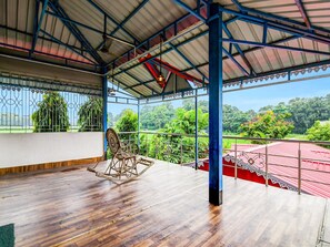 A covered outdoor area with a wooden floor, a rocking chair, metal railings, and a view of greenery. - Classic With Balconymoner Manush (Alipurduar)