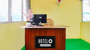 Un mostrador de recepción de hotel con un cartel que dice 'HOTELO Company-Serviced', una computadora, un ventilador y un jarrón con flores.